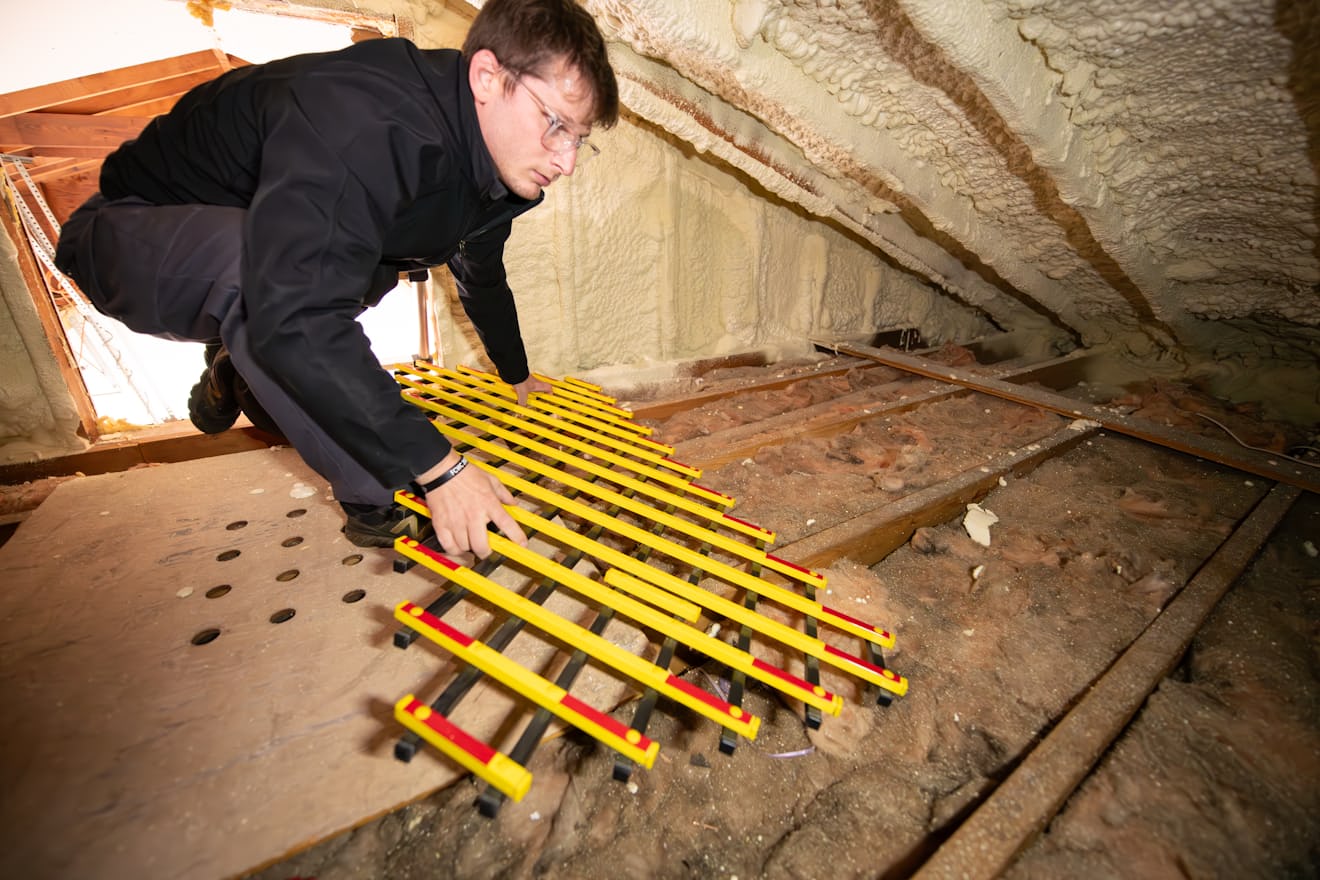 Worker kneeling on the floor of a residential attic beside insulation and roof framing